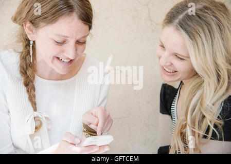 Zwei lächelnde Frauen mit langen blonden Haaren sitzen an einem Tisch, Blick auf ein Handy Stockfoto