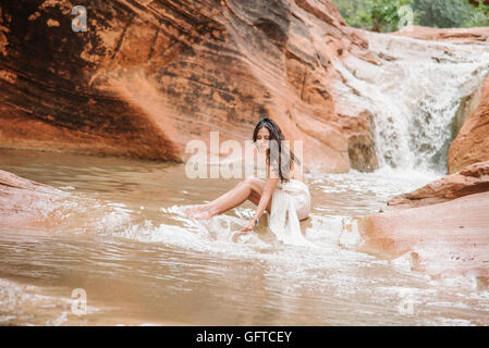 Junge Frau mit langen braunen Haaren, trägt ein langes weißes Kleid sitzt auf einem Felsen in einem Fluss Stockfoto