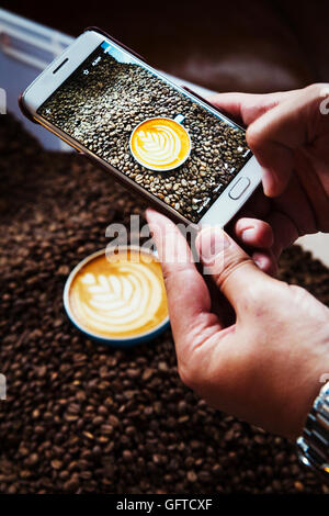 Eine Person, die ein Bild mit einem Smartphone für eine Tasse Kaffee auf einem Haufen von gerösteten Kaffeebohnen Stockfoto