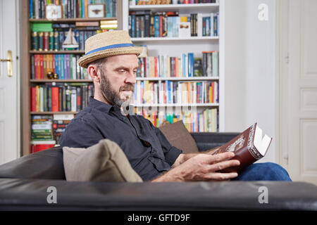 Student mit einem Buch vor einem Bücherregal auf einer Couch Hut Stockfoto