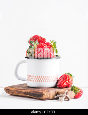 Frische reife rote Erdbeeren im Land Stil Emaille Becher auf rustikalem Holzbrett auf weißem Hintergrund Stockfoto