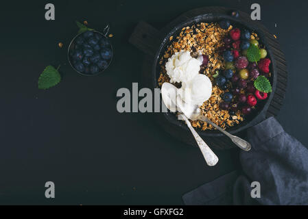 Gesundes Frühstück. Hafer-Müsli Crumble mit frischen Beeren, Samen und Eis in Bratpfanne Eisenpfanne auf dunklen Holzbrett auf schwarzem Hintergrund Stockfoto