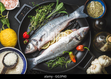 Zutaten für das Kochen gesunde Fisch essen. Rohe ungekochte Seebarsch mit Reis, Zitrone, Kräutern und Gewürzen auf schwarzes Grillen Eisen Pfanne auf dunklem Hintergrund Stockfoto