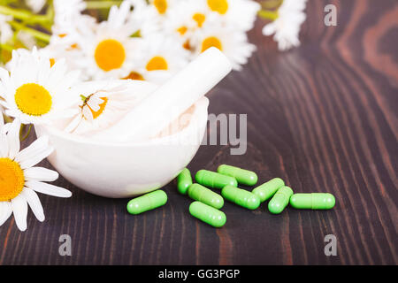 Kamillenblüten und grüne phytotherapeutische Kapseln auf einem Holztisch. Pflanzliche Arzneimittel und Gesundheit Stockfoto