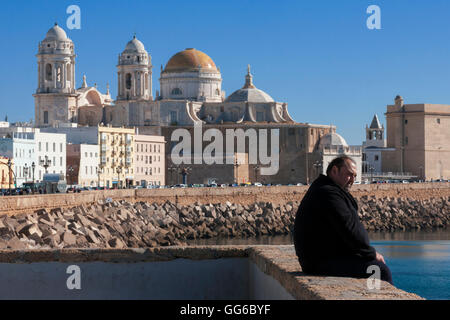 Mann sitzt an der Wand mit Blick auf den Ozean, mit dem Dom im Hintergrund: Calle Campo del Sur, Cádiz, Andalusien, Spanien Stockfoto