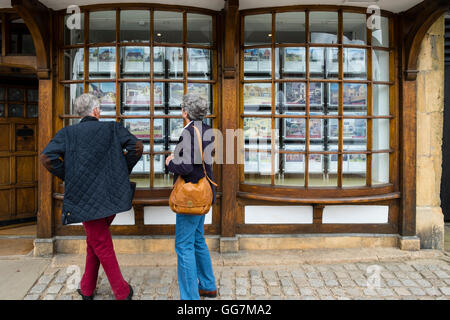 Älteres Paar sucht in Immobilienmakler Fenster an Immobilien zum Verkauf in den Cotswolds im England, United Kingdom Stockfoto