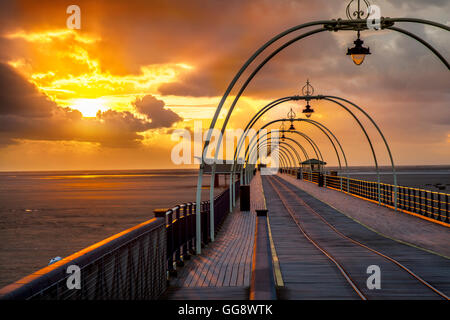 Southport, Merseyside, England. 9. August 2016. Dunkle Gewitterwolken über der veteran Pier Struktur nach Regenschauer, kühlt die Luft, kurz vor Sonnenuntergang.  Die tief rote Kugel der untergehenden Sonne richtet sich fast mit der Reflexion auf die Straßenbahn-Linien schafft eine Illusion einer Resort-Reise in die Unendlichkeit irgendwo heraus über die irische See Küste mit den Abend Strahlen Rötung Eisenkonstruktion. Bildnachweis: MediaWorldImages/Alamy Live-Nachrichten Stockfoto