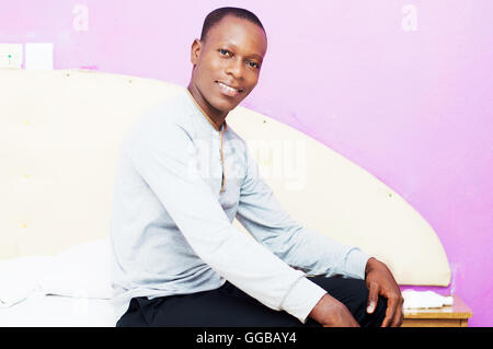junger Mann gekleidet am Morgen sitzt auf seinem Bett fröhlichen Gesicht zur Kamera. Stockfoto