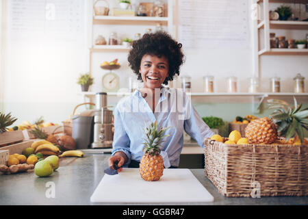 Porträt der fröhliche junge afrikanische Frau stehend hinter Theke mit Ananas auf Schneidebrett. Arbeiten bei Saft weiblich Stockfoto