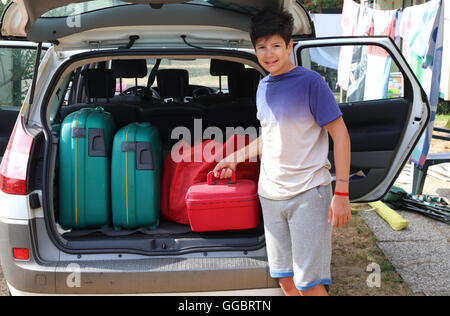 lächelnde junge lädt einen wenig roten Koffer in den Kofferraum des Autos vor seiner Abreise nach einer langen Reise Stockfoto