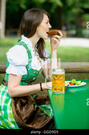 junge Frau im Dindl am Biergarten im Freien sitzen und essen Brot Stockfoto