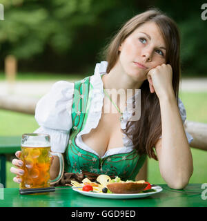 junge Frau im Dirndl mit Essen und Bier im Biergarten sitzen und schauen gelangweilt Stockfoto