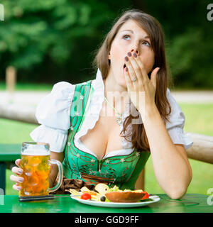 junge Frau im Dirndl sitzen in Bier Garten mit Essen und Bier und ist Gähnen Stockfoto