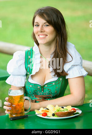 junge bayerische Frau im Dirndl mit Bier und Essen am Tisch im Biergarten sitzen Stockfoto