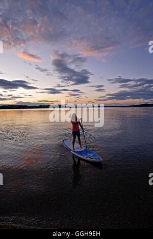 Frau Stand-up Paddeln auf einem See Chiemsee im Sonnenuntergang, Chiemgau, Bayern, Deutschland Stockfoto