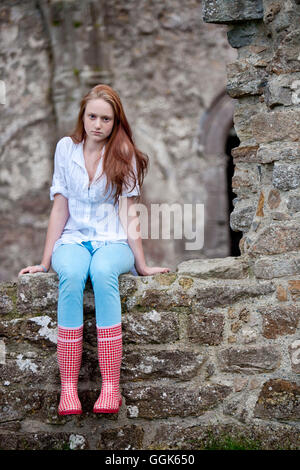 Rote behaarter Teenager-Mädchen tragen Wellington boots mit roten Punkten sitzen auf einer Steinmauer, Clonmacnoise, County Offaly, Irland, Eu Stockfoto