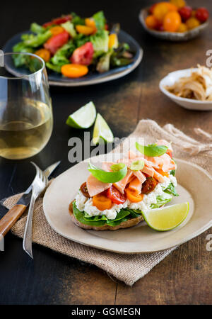 Sandwich mit Frischkäse, Lachs und Tomaten auf Teller über rustikalen hölzernen Hintergrund, Weißwein und frischem Salat auf Seite, selec Stockfoto