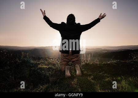 Heckansicht des ein Mann mit ausgestreckten Armen, kniet in der Wiese auf einem Hügel. Stockfoto