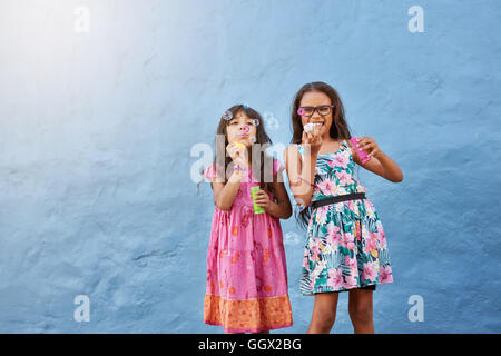 Porträt von niedlichen kleinen Mädchen bläst Seifenblasen. Zwei junge Mädchen spielen vor blauem Hintergrund. Stockfoto