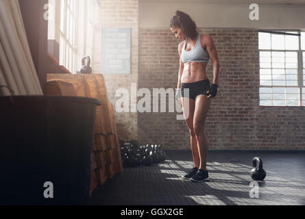 Eine lange Aufnahme der Passform und der jungen Frau, die im Crossfit-Fitnessstudio steht. Fitnessmodell für Frauen mit Kesselglocke auf dem Boden. Stockfoto