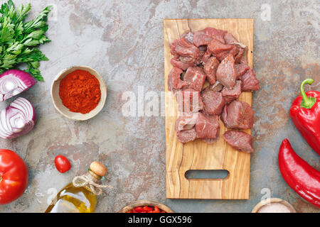 Rindfleisch für Gulasch. Fleischwürfel auf einem alten hölzernen Schneidebrett und Zutaten für das Kochen auf rustikalen Hintergrund Stockfoto