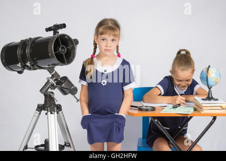 Eine Mädchen steht ein Teleskop mit seinen Händen in den Hosentaschen, ein anderes Mädchen Spaß sitzen am Tisch und schreibt Stockfoto