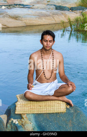 Ein junger Hindu Priester (Brahmanen) sitzt in der Meditation am Ufer eines Flusses. Stockfoto