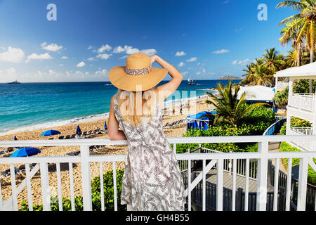 Rückseite Blick auf eine Frau in einem Sommerkleid und Hut ein Beach Resort von einem Balkon aus auf Stockfoto