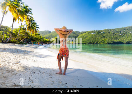 Frau im Sarong und Hut stehend an einem tropischen Strand mit beiden Händen hinter ihr, Magens Bay, St. Thomas, Amerikanische Jungferninseln Stockfoto