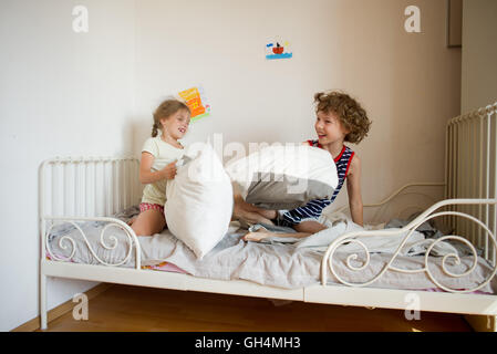 Kleinen Bruder und Schwester inszeniert eine Kissenschlacht im Schlafzimmer. Sie sitzen auf einem Bett im Pyjama. Kinder Haare ungekämmt. Glücklich smil Stockfoto
