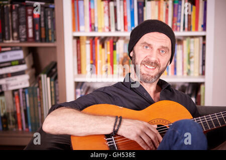 Hipster-Mann mit Bart und Hut sitzt auf einer Couch vor einem Bücherregal Gitarre spielen Stockfoto