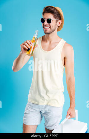 Fröhlichen jungen Mann in Hut und eine Sonnenbrille Kühltasche halten und trinken Bier Stockfoto