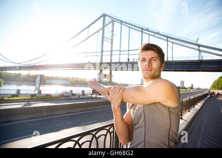 Hübscher Junge Sport Mann tut erstreckt sich auf das Brückengeländer und wegschauen im freien Stockfoto