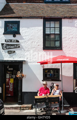 England, Broadstairs. Einer langen Ehe; ältere kaukasischen paar sitzt unter Sonnenschirm Regenschirm, außerhalb der touristischen Attraktion, "The Old Curiosity" Shop. Mann und Frau auf der Suche von einander entfernt. Sommer, Sonnenschein. Stockfoto