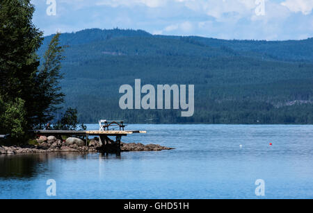 Holzbrücke, Tisch, Stühlen und einer linken Angelrute. See und Grate, Wälder in der Umgebung. Sommer. Stockfoto