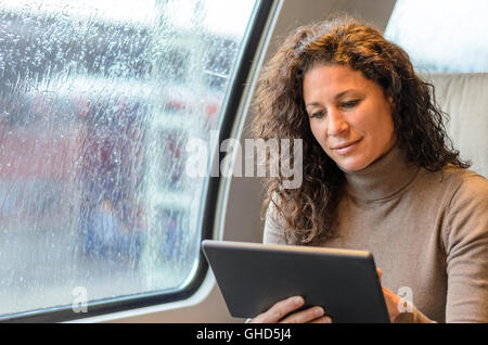 Junge Frau, die das Surfen im Internet während der Fahrt in einem Zug an einem regnerischen Tag mit einem Handheld-Tablet mit einem ruhigen Lächeln Stockfoto