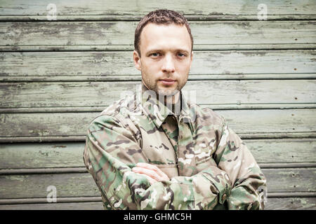 Ernsthafte kaukasischen militärischen Jüngling in Camouflage uniform mit Händen überqueren. Closeup Portrait über grüne Holzwand Stockfoto