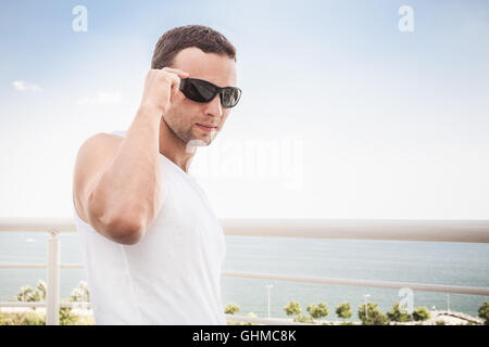 Junge sportliche lächelnder kaukasischen Mann in weißem Hemd und Sonnenbrille. Outdoor-Sommer-Porträt mit Meerblick auf einem Hintergrund Stockfoto