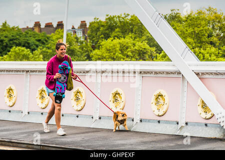 Eine junge Frau, die mit einem Hund auf einem Skateboard auf der Albert Bridge, Chelsea, London, England, Großbritannien, läuft Stockfoto