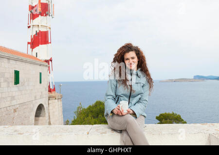 Junge Frau nimmt sich eine Auszeit mit Leuchtturm im Hintergrund Stockfoto