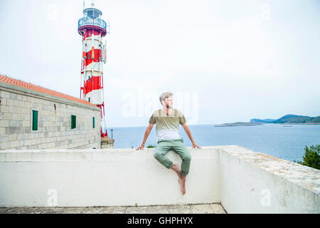 Junger Mann sitzt auf Wand mit Leuchtturm im Hintergrund Stockfoto