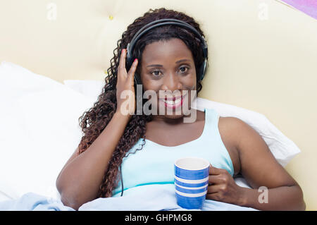 junge Frau im Bett liegend freut sich aufwachen, um Musik und einer Tasse Kaffee in der hand Stockfoto