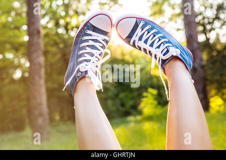 Blick auf Frau Beine tragen Jeans Turnschuhe über einen Sonnenuntergang Strahlen in einem Wald in der Nähe Stockfoto