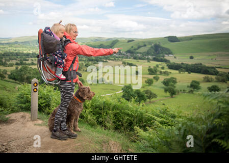 Eine junge Frau mit Baby im Tragetuch zeigt auf etwas in der Landschaft wandern. Wandern mit Kind und Hund o Stockfoto