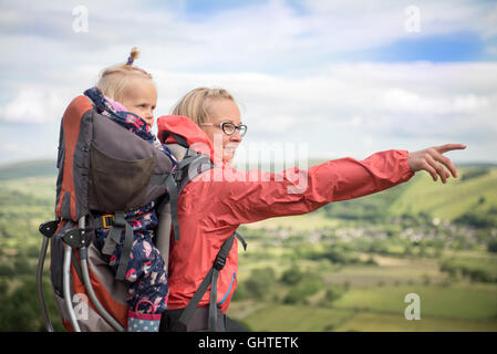 Glückliche junge Frau trekking mit Baby im Tragetuch und ihr einen Hund zeigte auf etwas in der Landschaft. Wandern Aktivität w Stockfoto