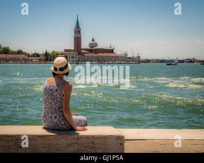 Rückansicht der weiblich, mit Stroh Hut, sitzt auf einer Bank Blick auf die Kirche und die Insel San Giorgio Maggiore. Stockfoto