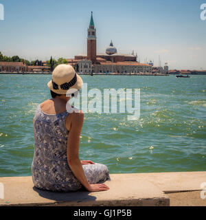 Rückansicht des einzigen Weibchen, mit Stroh Hut, sitzt auf einer Bank Blick auf die Kirche und die Insel San Giorgio Maggi Stockfoto