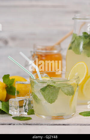 Glas Limonade mit Zitronenscheibe, Minze Blätter und Eiswürfel in der Nähe von Zitronen Limonade Flasche und Honig jar auf rustikale weißen Holz Stockfoto