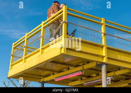 Ein junges Paar, küssen, blind für Passanten, stehend auf einer gelben Plattform an einem sonnigen Tag, erhaben über dem Bürgersteig. Stockfoto