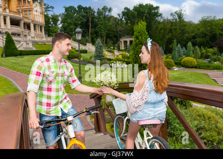 Schöner junger Mann gibt einer Mädchen ein Bouquet von kleinen weißen Blumen als Geschenk, während sie auf dem Fahrrad am Steg im Park unterwegs sind. Stockfoto